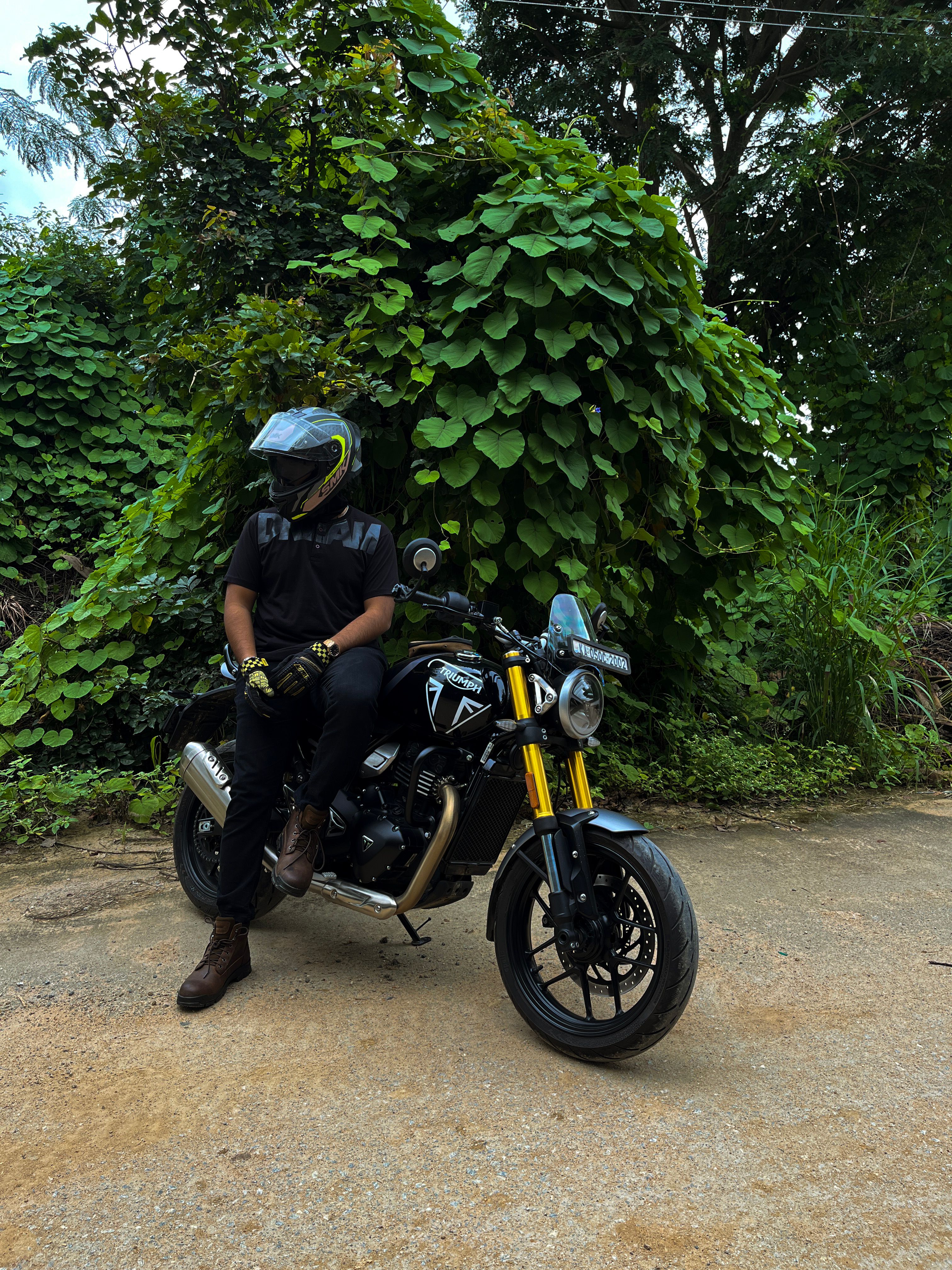 A helmeted rider sits on a Triumph bike against a lush, green backdrop.