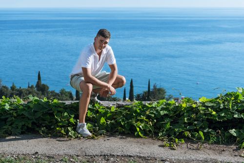 Free A young man crouching by the ocean, surrounded by greenery, enjoying a sunny day outdoors. Stock Photo