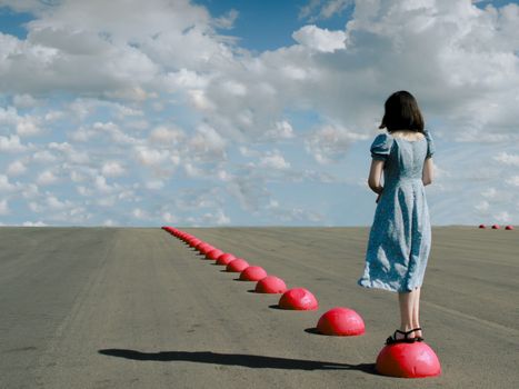 A young woman in a blue dress stands on red spheres aligned on an open road.