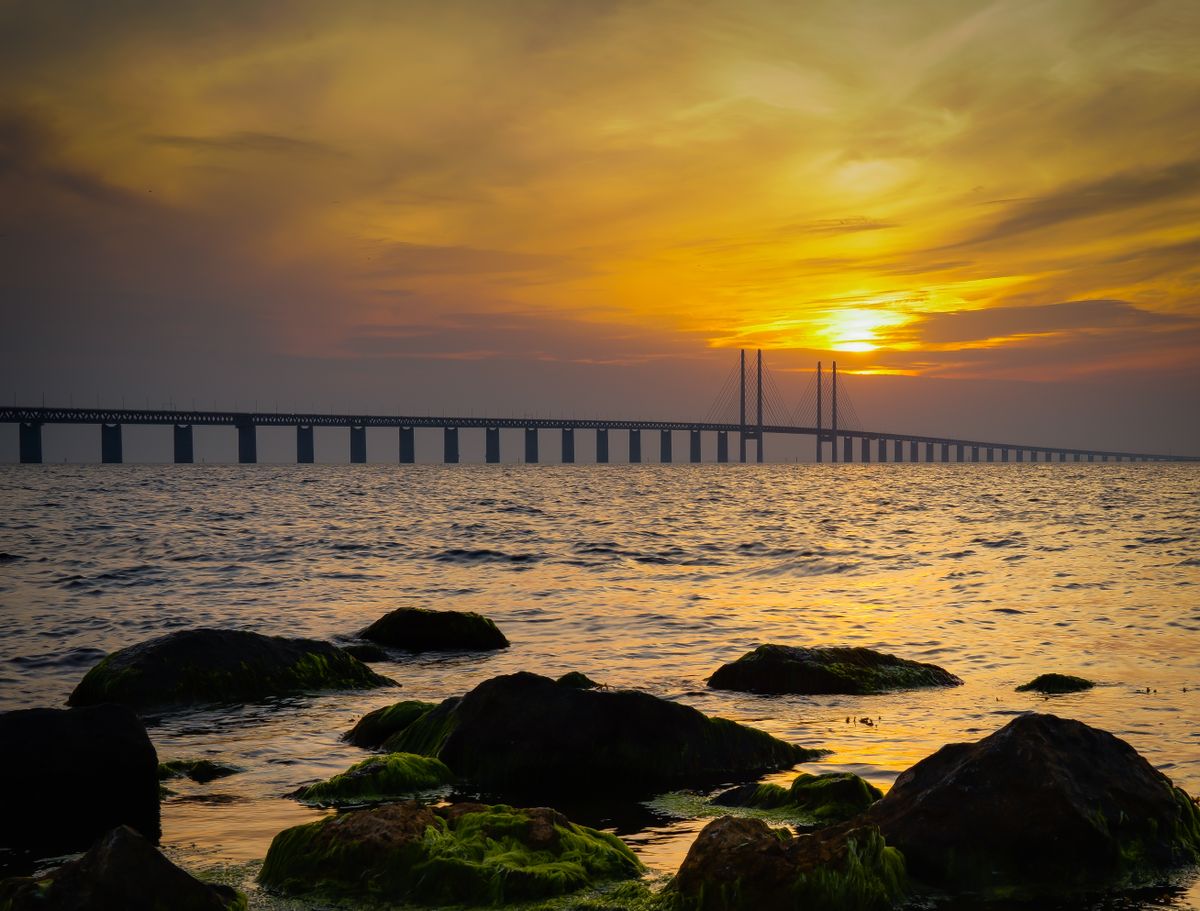 Podul Øresund întinzându-se spre orizont la apus, văzut de pe un țărm cu bolovani