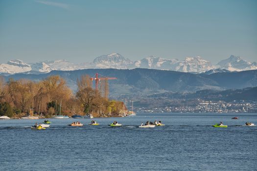 A picturesque view of Lake Zurich with people boating and the snowy Alps in the background.