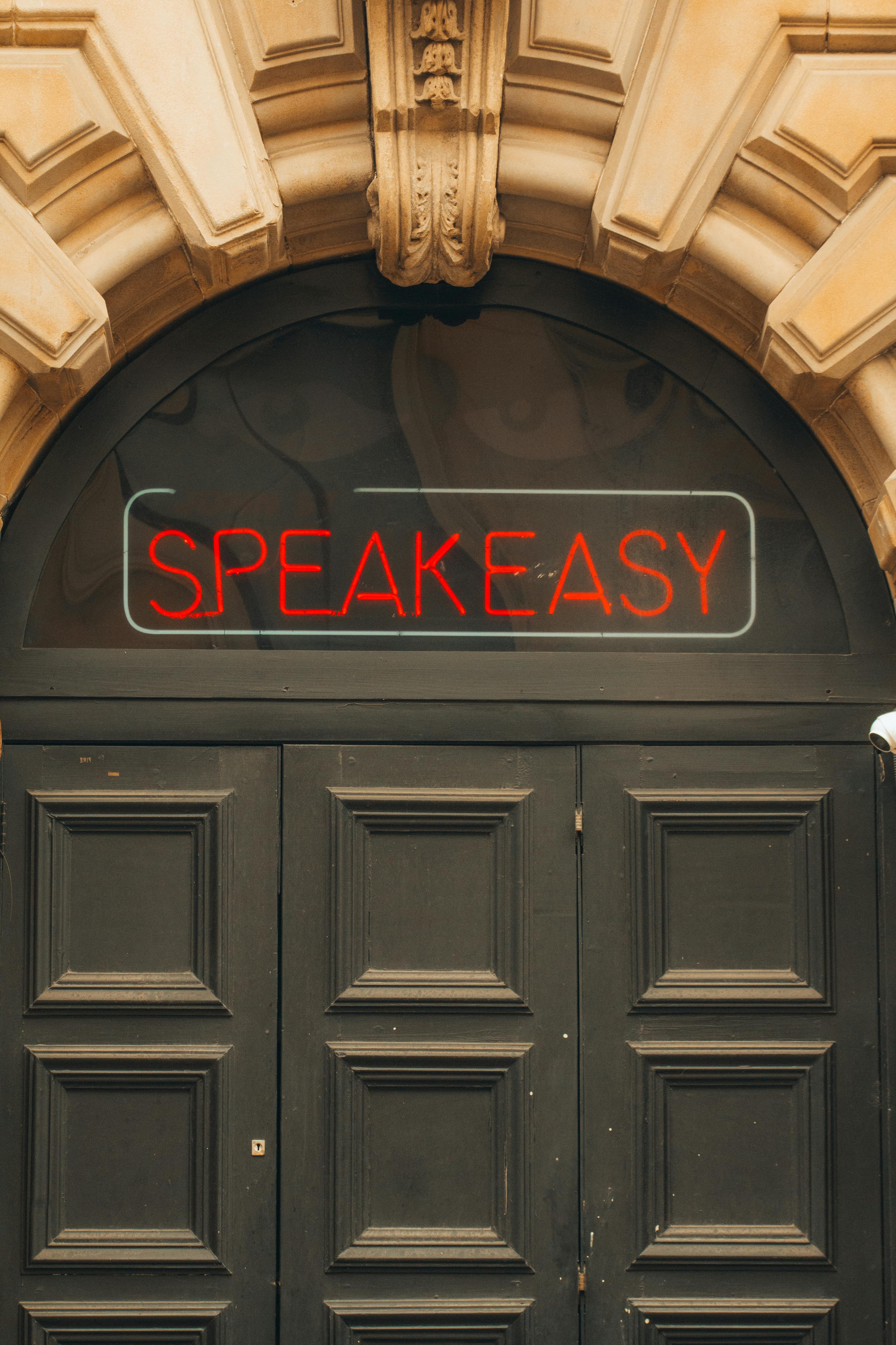 Close-up of an ornate wooden door with a glowing speakeasy sign above.