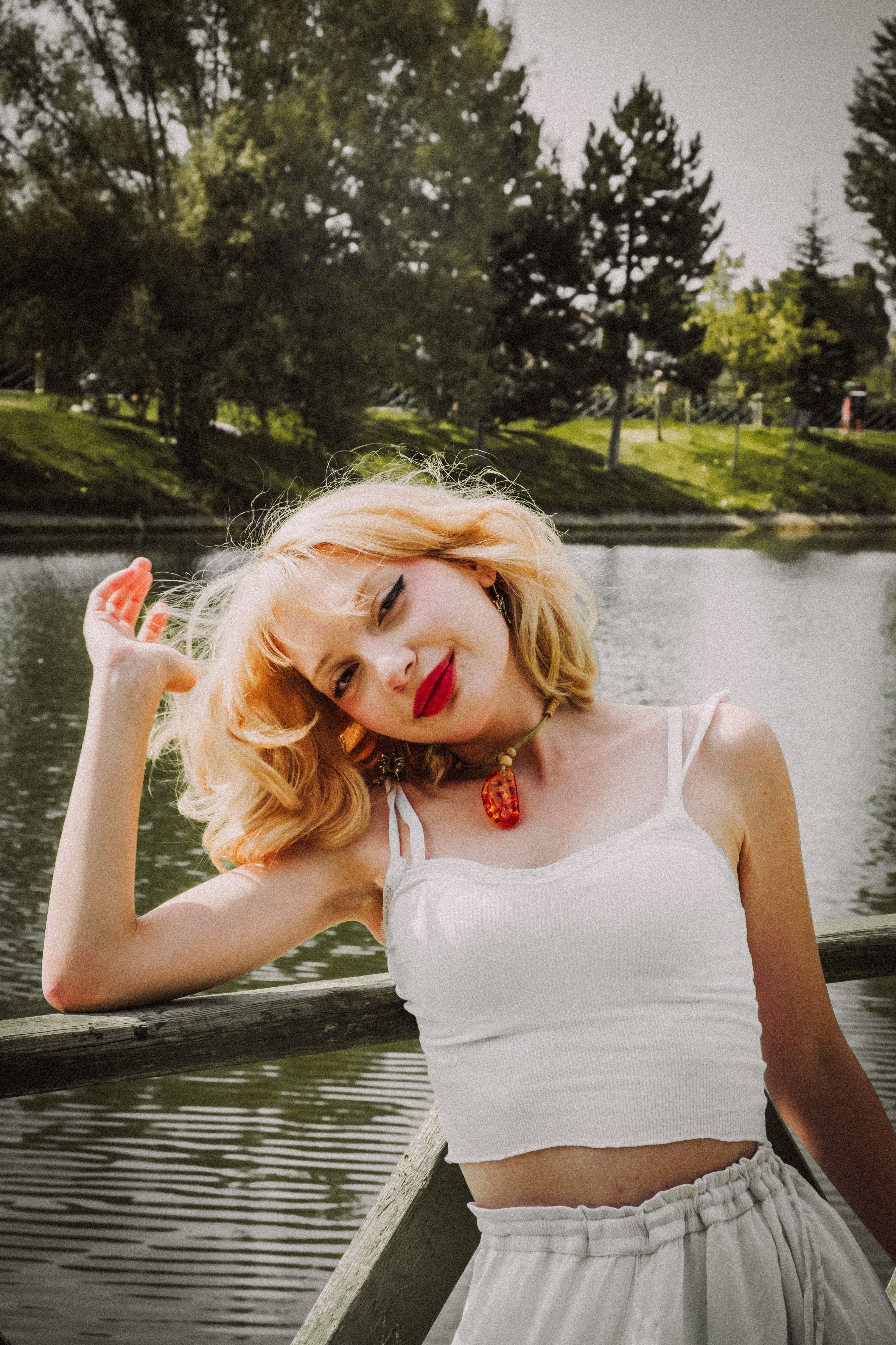 A blonde woman enjoying a sunny day by the lake, wearing a white dress and red lipstick.