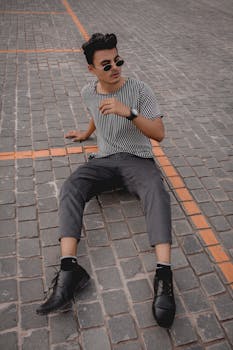 一位戴着墨镜的时尚年轻人以休闲的姿势坐在铺好的街道上，展现出一种现代风格