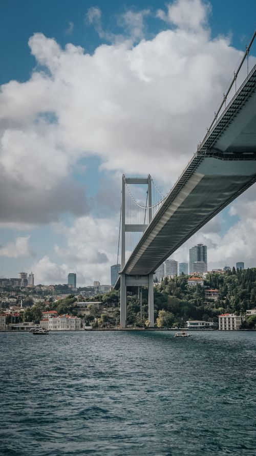 Free A modern suspension bridge spanning over the water with a vibrant cityscape in the background. Stock Photo