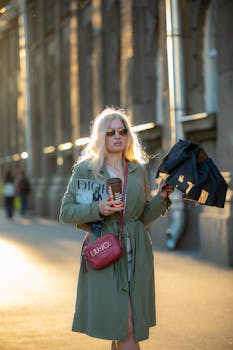 Fashionable woman with umbrella and coffee walking in urban area at sunset.