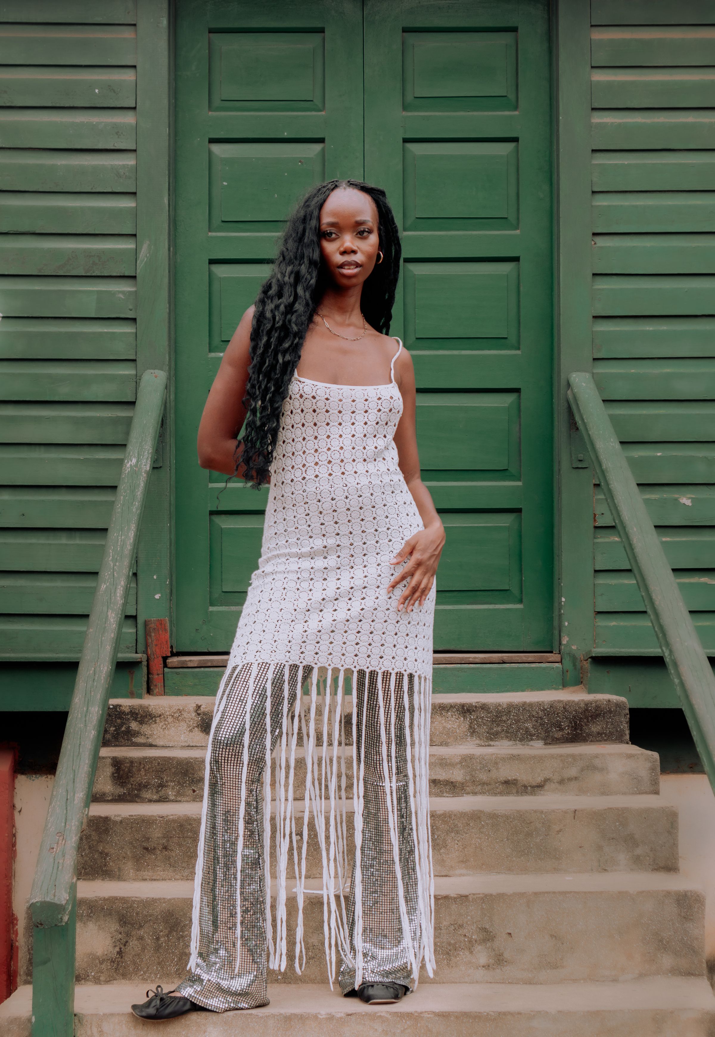 A Ghanaian woman in a white dress stands on the steps of a green wooden house in Accra.