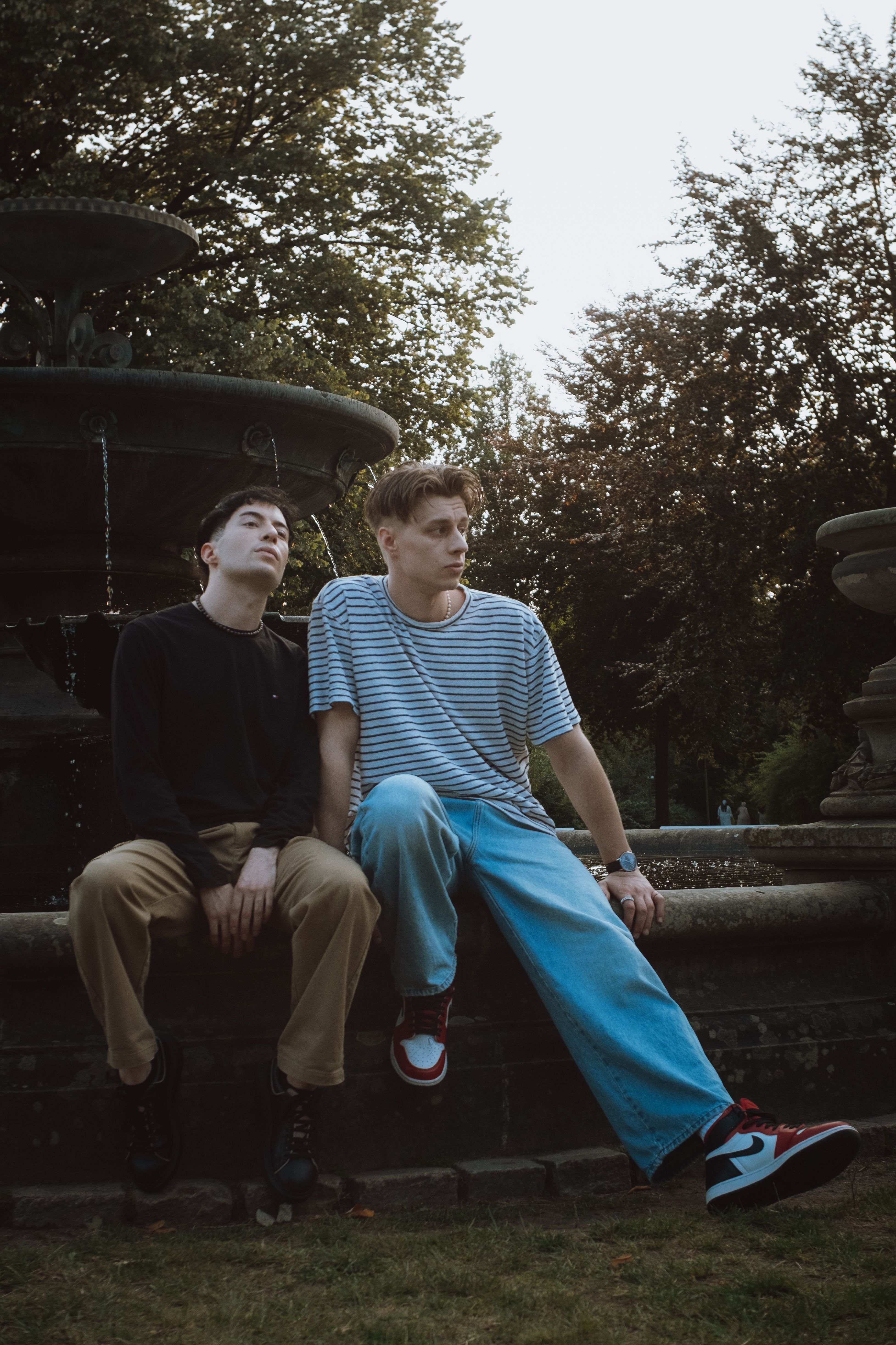 Two young men sitting by a fountain in a park, enjoying a relaxed moment outdoors.