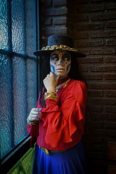 Woman in Day of the Dead costume with makeup and hat indoors near brick wall.