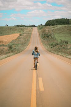 一位金发女孩骑着自行车，沿着风景秀丽的长长高速公路，穿行在乡村田野间