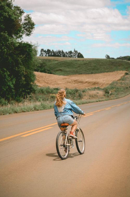 Free A woman with curly hair rides a bicycle on a quiet country road surrounded by lush greenery and open fields on a sunny day. Stock Photo