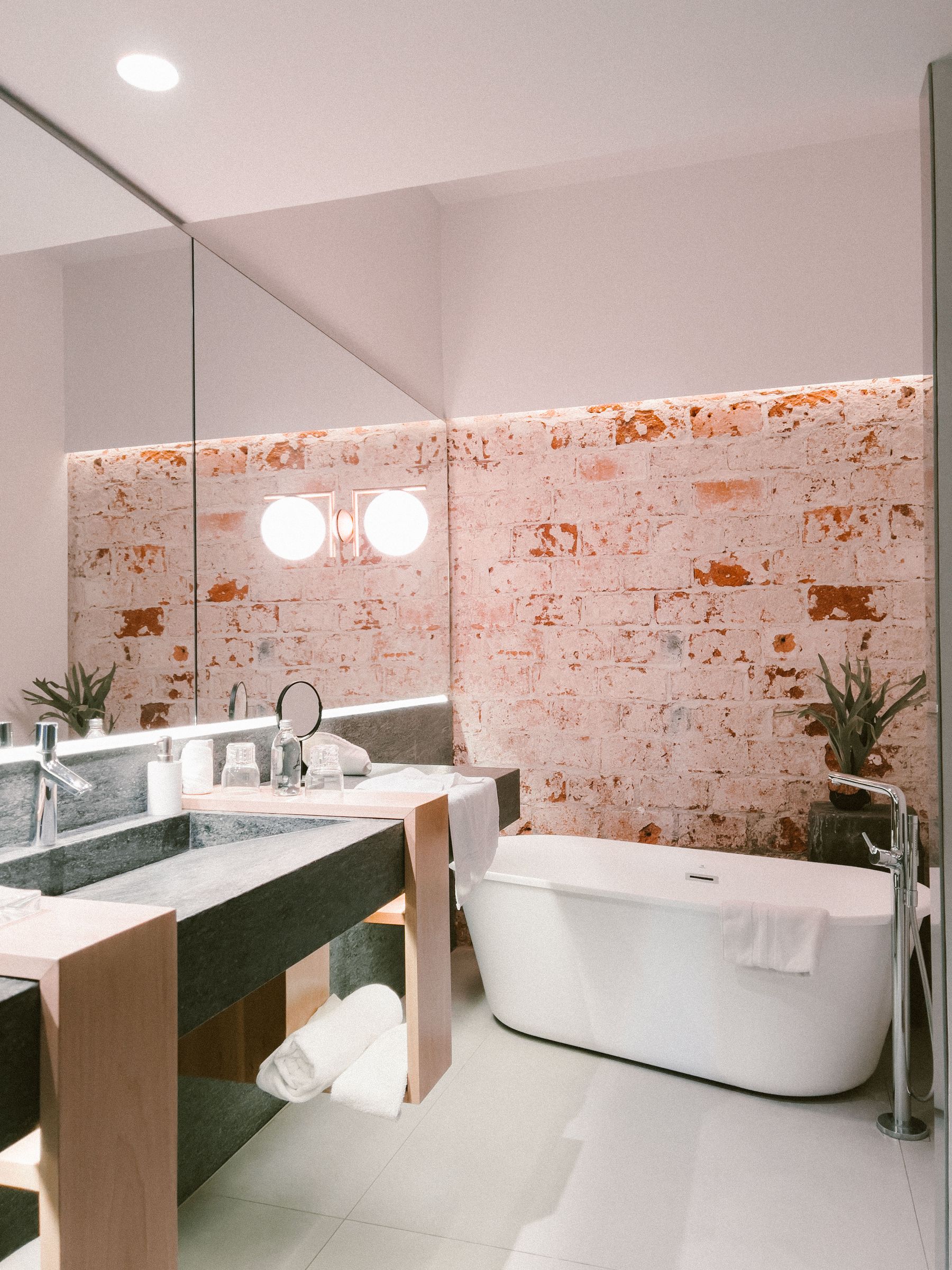 Industrial bathroom with exposed brick, a freestanding white bath, dark stone vanity and warm wood framing