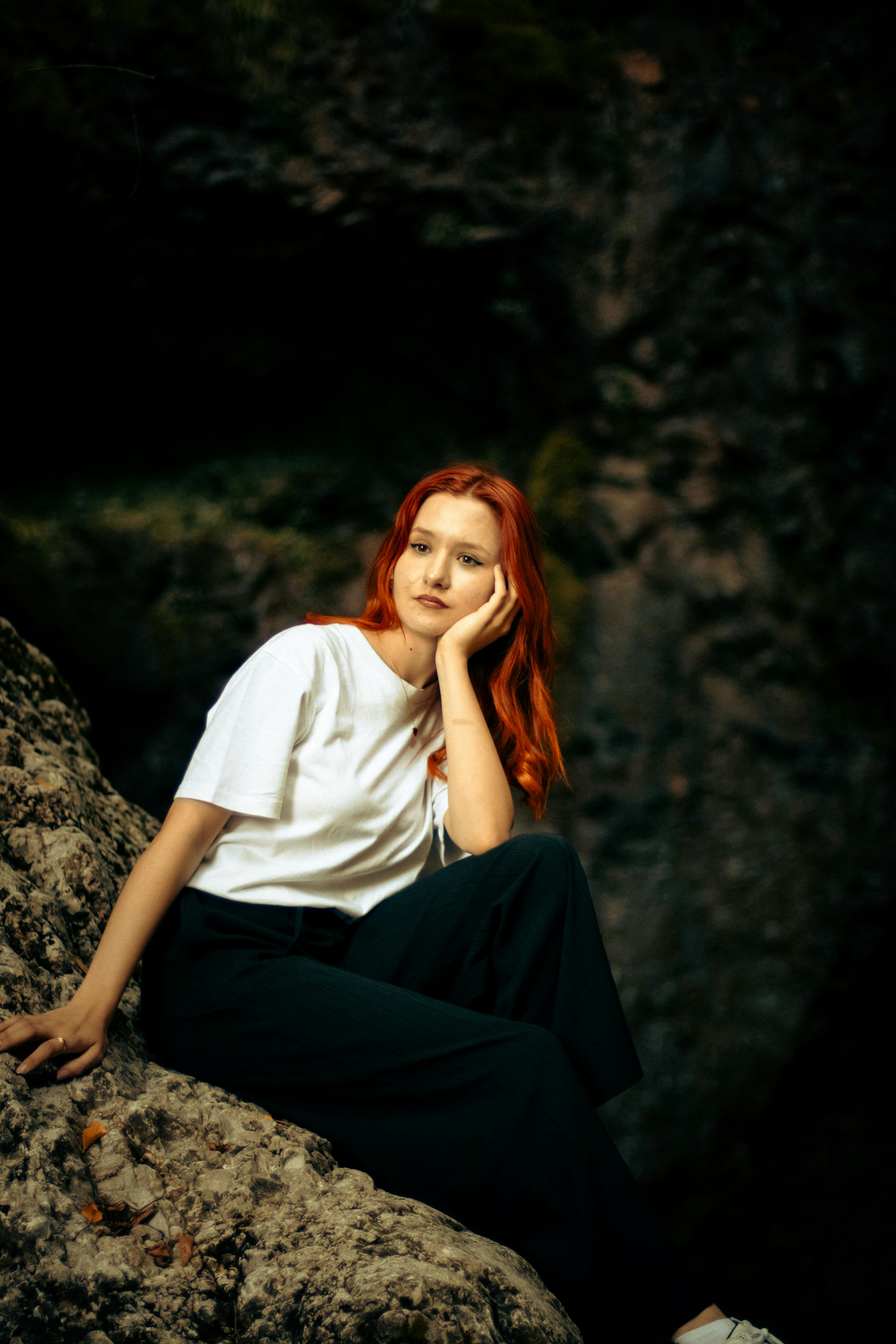 A pensive woman with red hair sits outdoors in a natural rocky setting, expressing relaxation.