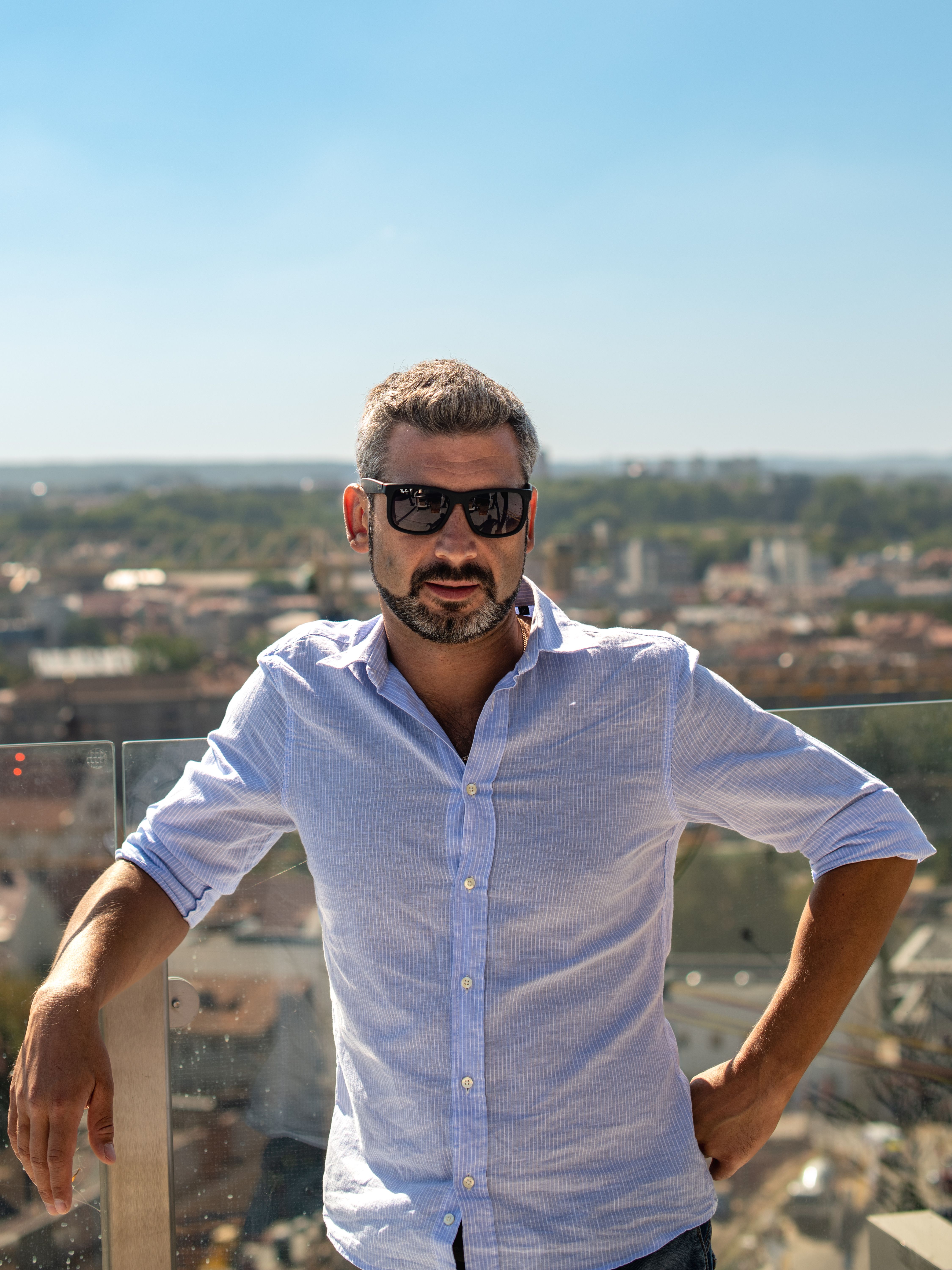 Confident man in sunglasses stands on a rooftop overlooking Vilnius skyline under the bright summer sun.
