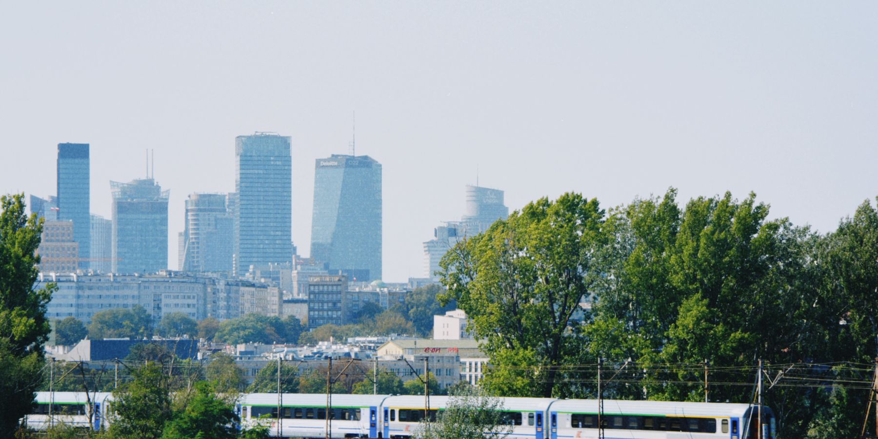 Skyline of Warsaw with modern buildings and a train passing amidst the greenery.