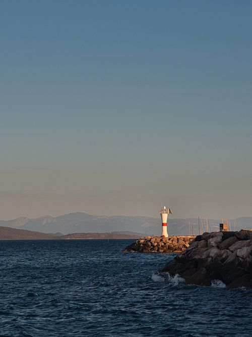 Free A peaceful view of a lighthouse on a rocky coast at sunset with mountains in the background. Stock Photo