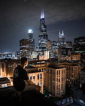 A man sits on a rooftop, overlooking the illuminated Chicago skyline at night.