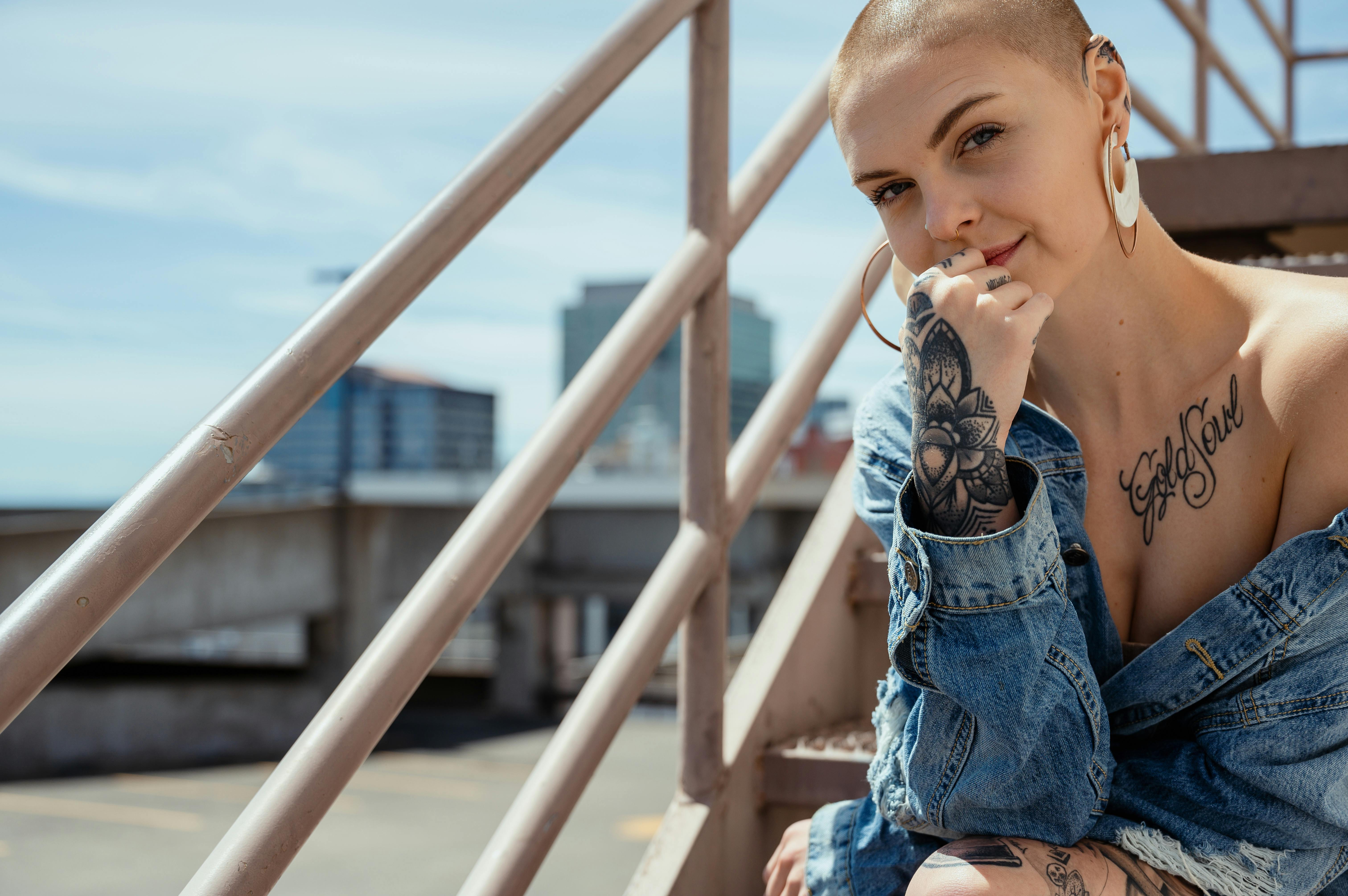 一位剃着光头、身穿牛仔夹克的纹身女子，在户外楼梯上自信地摆着姿势