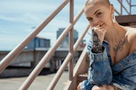 一位剃着光头、身穿牛仔夹克的纹身女子，在户外楼梯上自信地摆着姿势