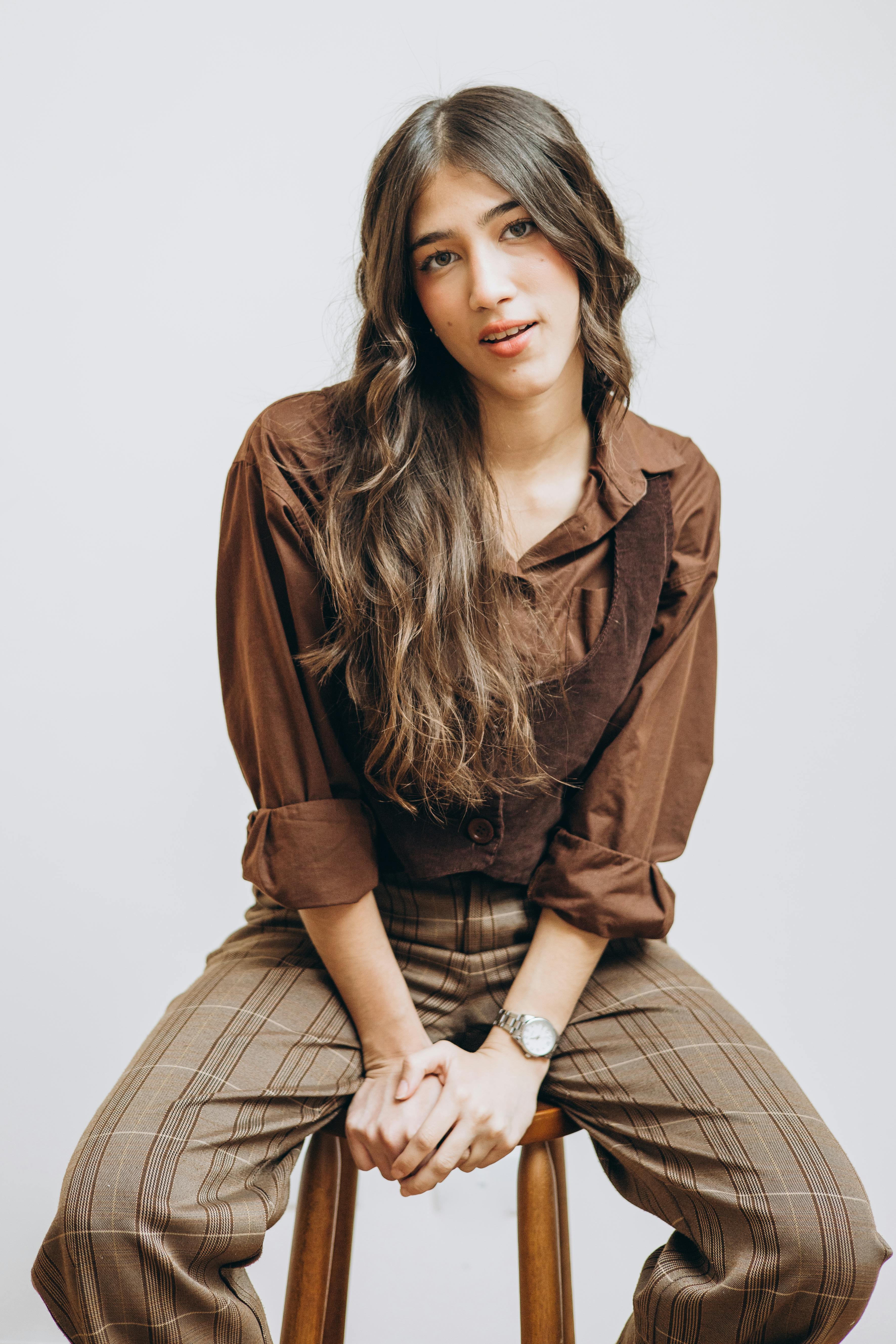 Chic woman in brown outfit posing on wooden stool in a modern studio setting, exuding confidence and style.