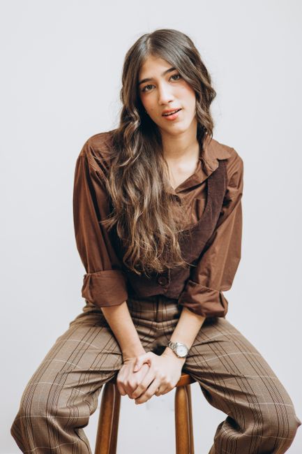 Chic woman in brown outfit posing on wooden stool in a modern studio setting, exuding confidence and style.