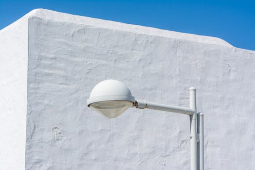 Minimalist white street lamp set against a textured white wall under a clear blue sky.