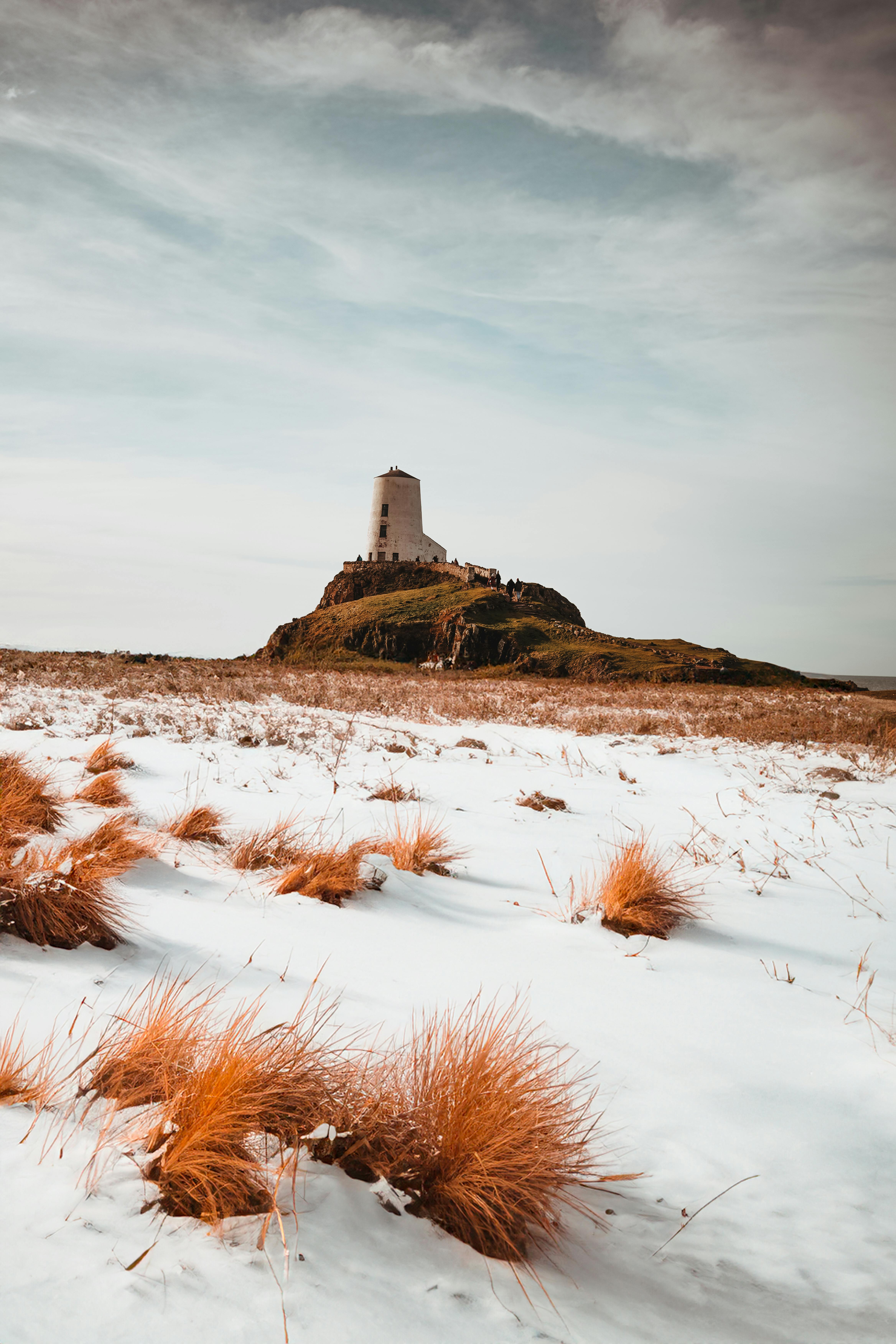 Scenic winter view of Ynys Llanddwyn lighthouse on a snowy island with grass tufts.