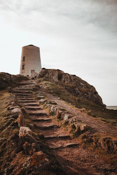 Scenic view of Ynys Llanddwyn lighthouse with stone stairs amidst rocky terrain under a cloudy sky.