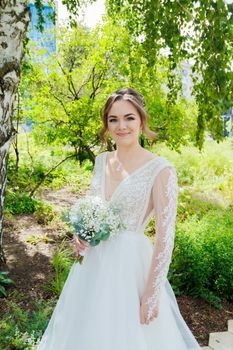 A stunning bride in a lace wedding dress holding a bouquet outdoors among greenery.