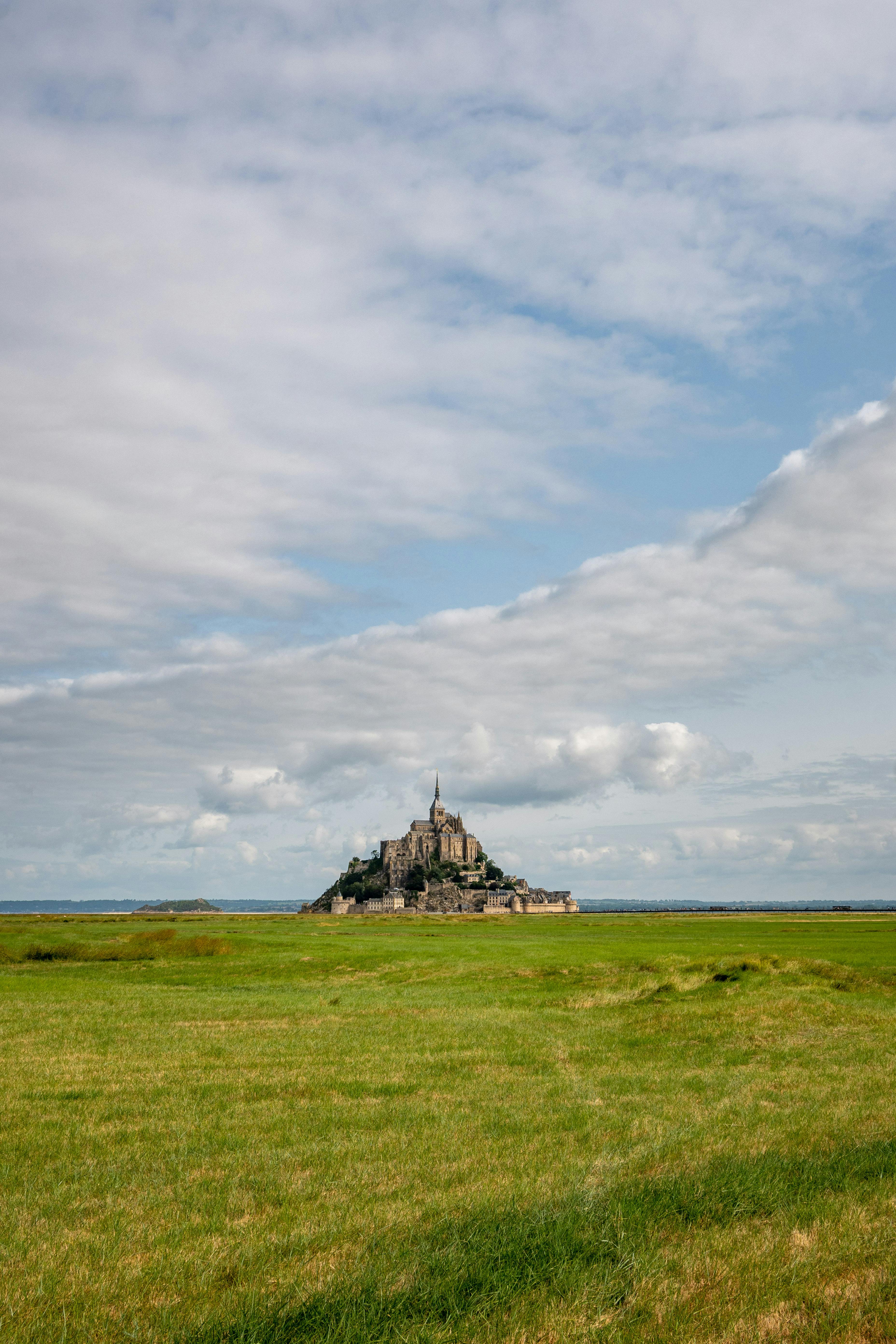 Scenic view of Mont Saint-Michel in Normandy, France with green fields under a partly cloudy sky.