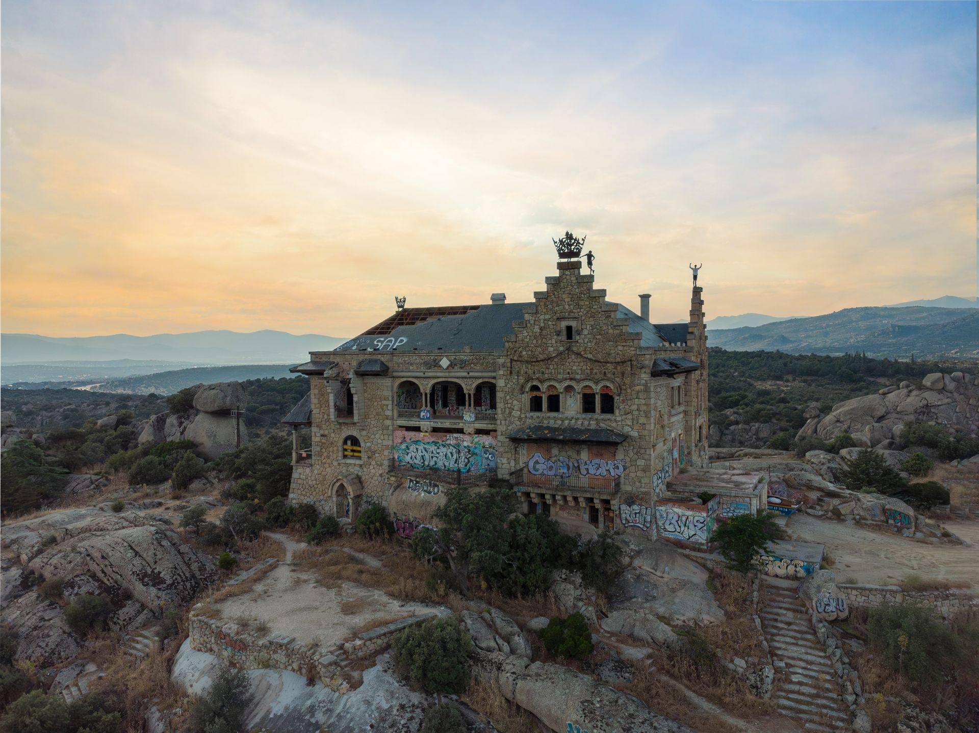 Aerial view of an abandoned mansion covered in graffiti on a rocky hillside at sunset.