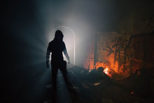 A silhouette of a hooded figure in a dark, moody tunnel with a glowing entrance and flames nearby.