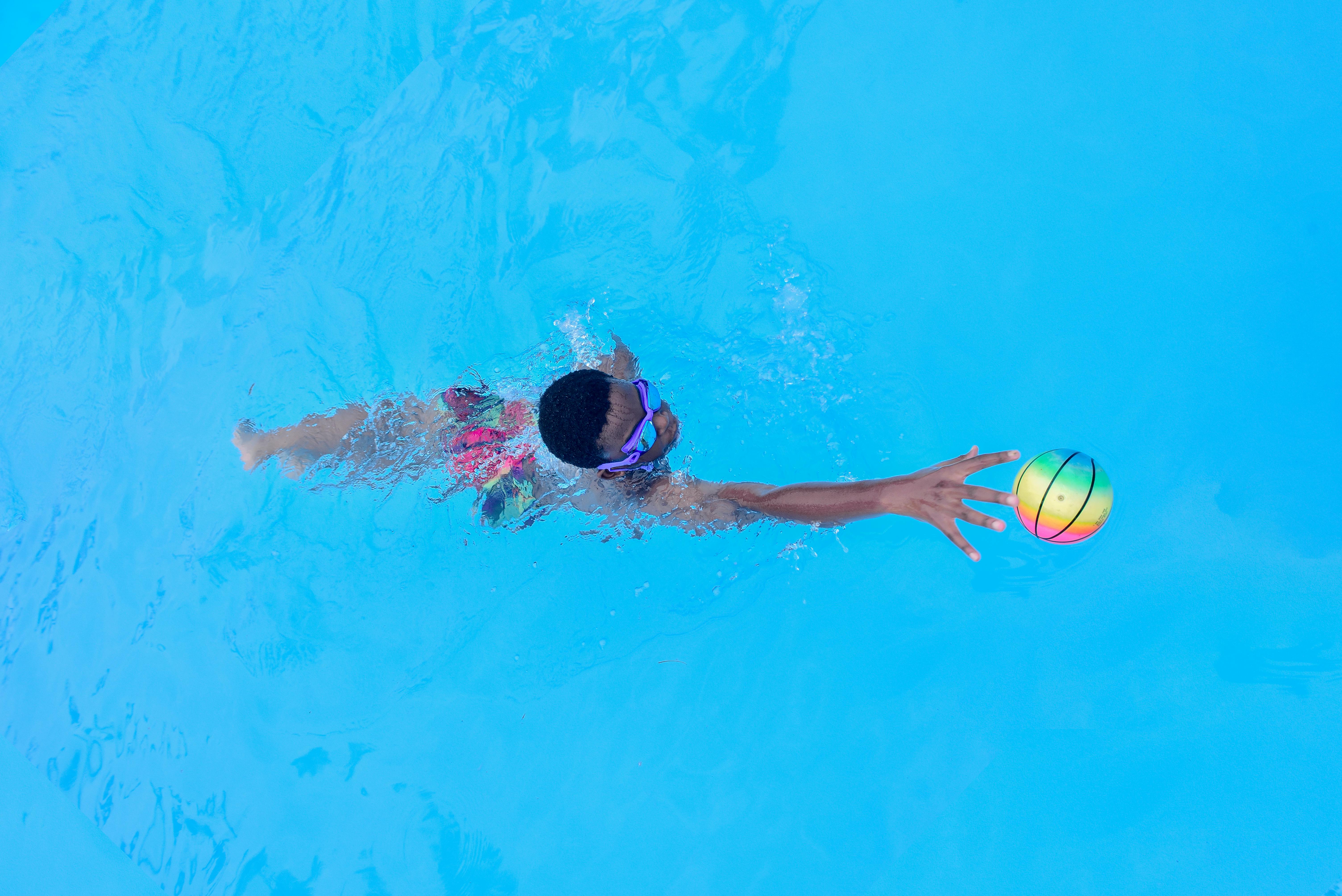 A child swimming underwater reaches for a colorful ball in a clear blue pool, enjoying a summer day.