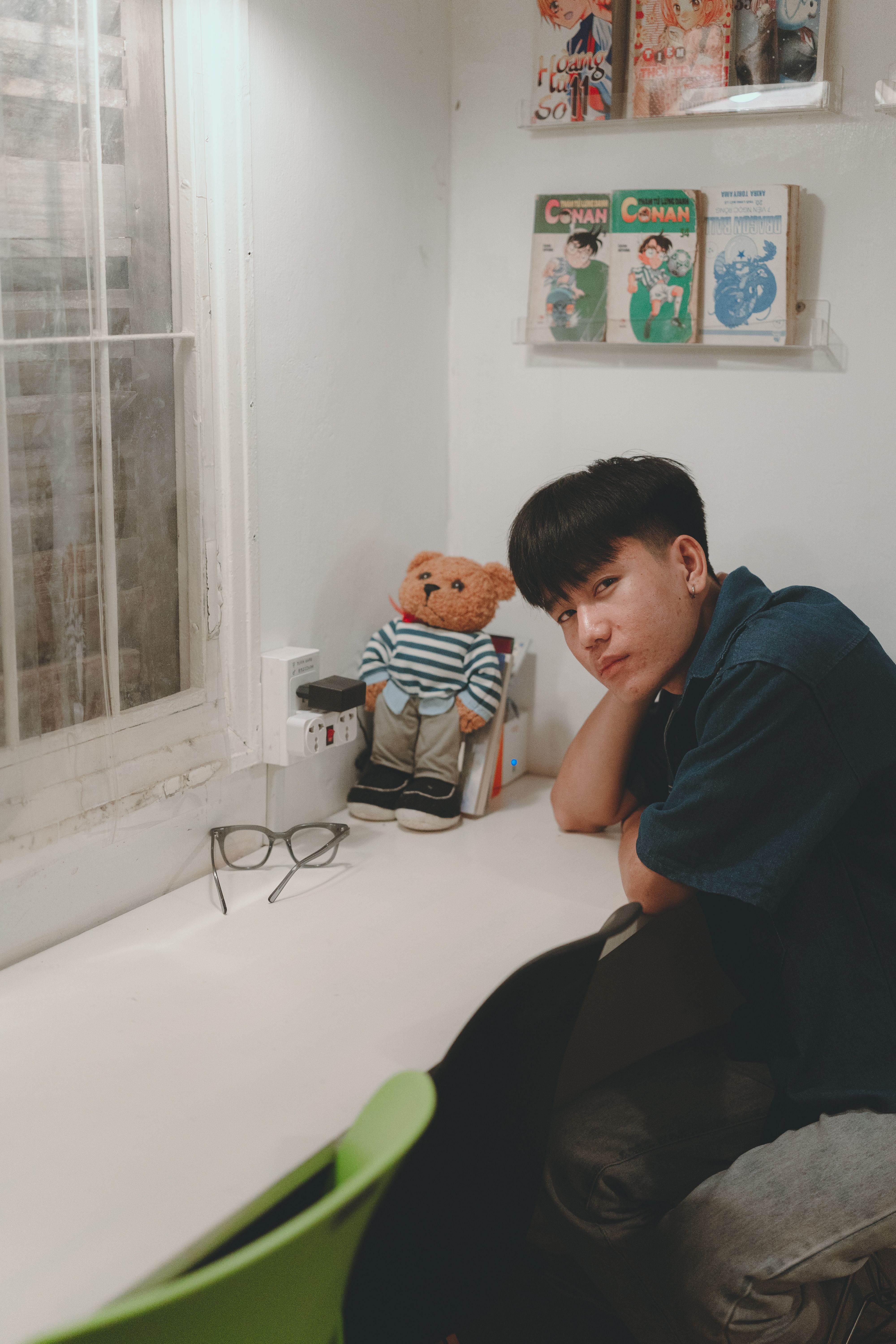 A young man relaxes at his desk indoors, accompanied by a teddy bear and comic books, creating a cozy atmosphere.