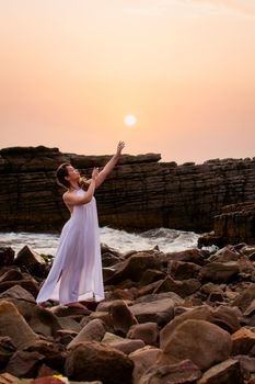 Graceful woman in white dress standing on rocky beach at sunset, embracing ocean beauty.