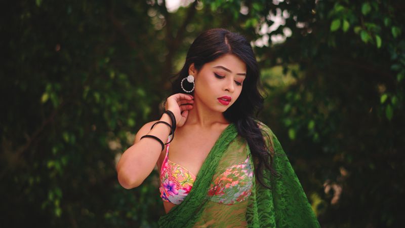 Young woman in a vibrant saree posed outdoors surrounded by lush greenery, exuding elegance and style.