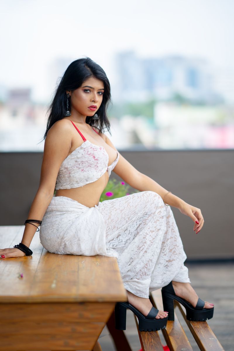 Confident woman in white lace saree sitting on a rustic table outdoors, exuding elegance and style.