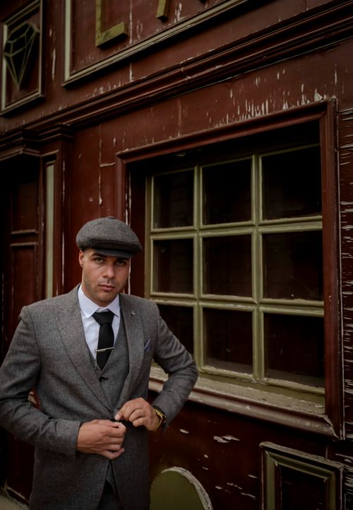Free A man in a classic suit and flat cap stands by a rustic building, exuding vintage charm. Stock Photo