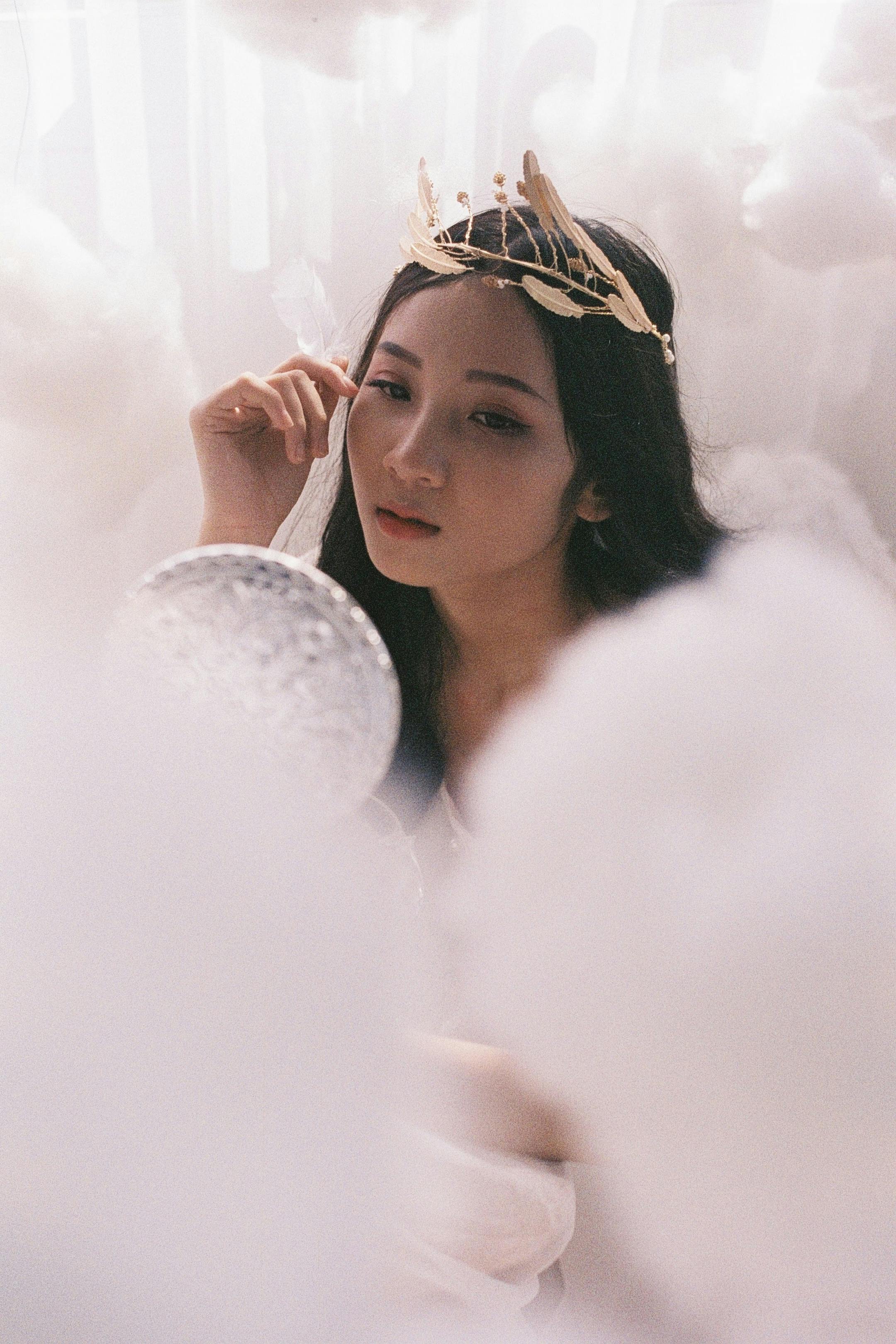 Ethereal portrait of an Asian woman with a crown, surrounded by clouds, holding a mirror.