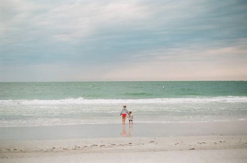 Free A woman and child enjoy a tranquil beach day, gazing at the ocean under an overcast sky. Stock Photo