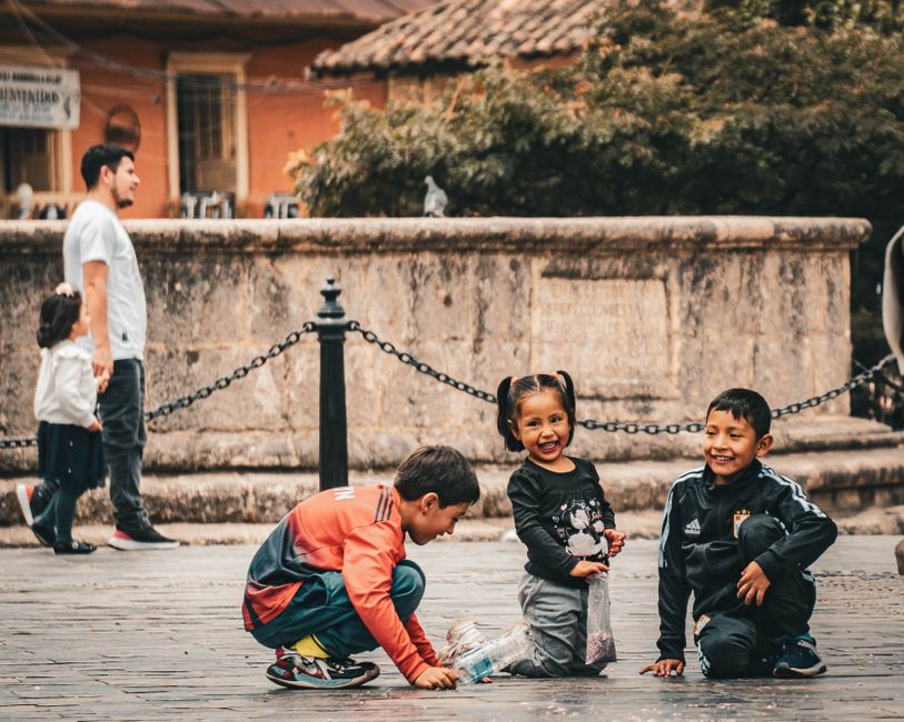 Tres niños juegan y sonríen en una animada plaza urbana, capturando una atmósfera vibrante de la ciudad.