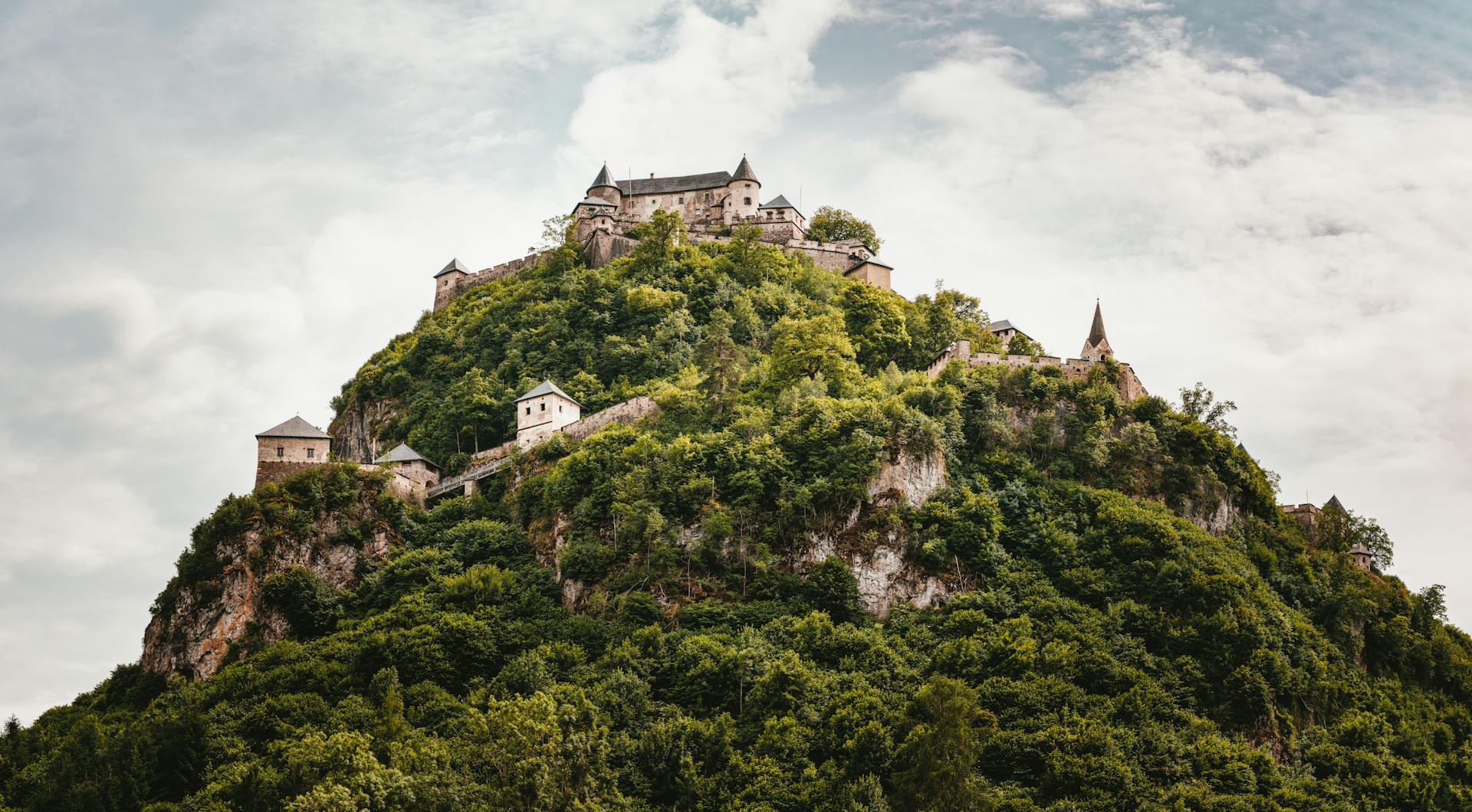 Beautiful medieval castle nestled atop a forested hill under a cloudy sky.