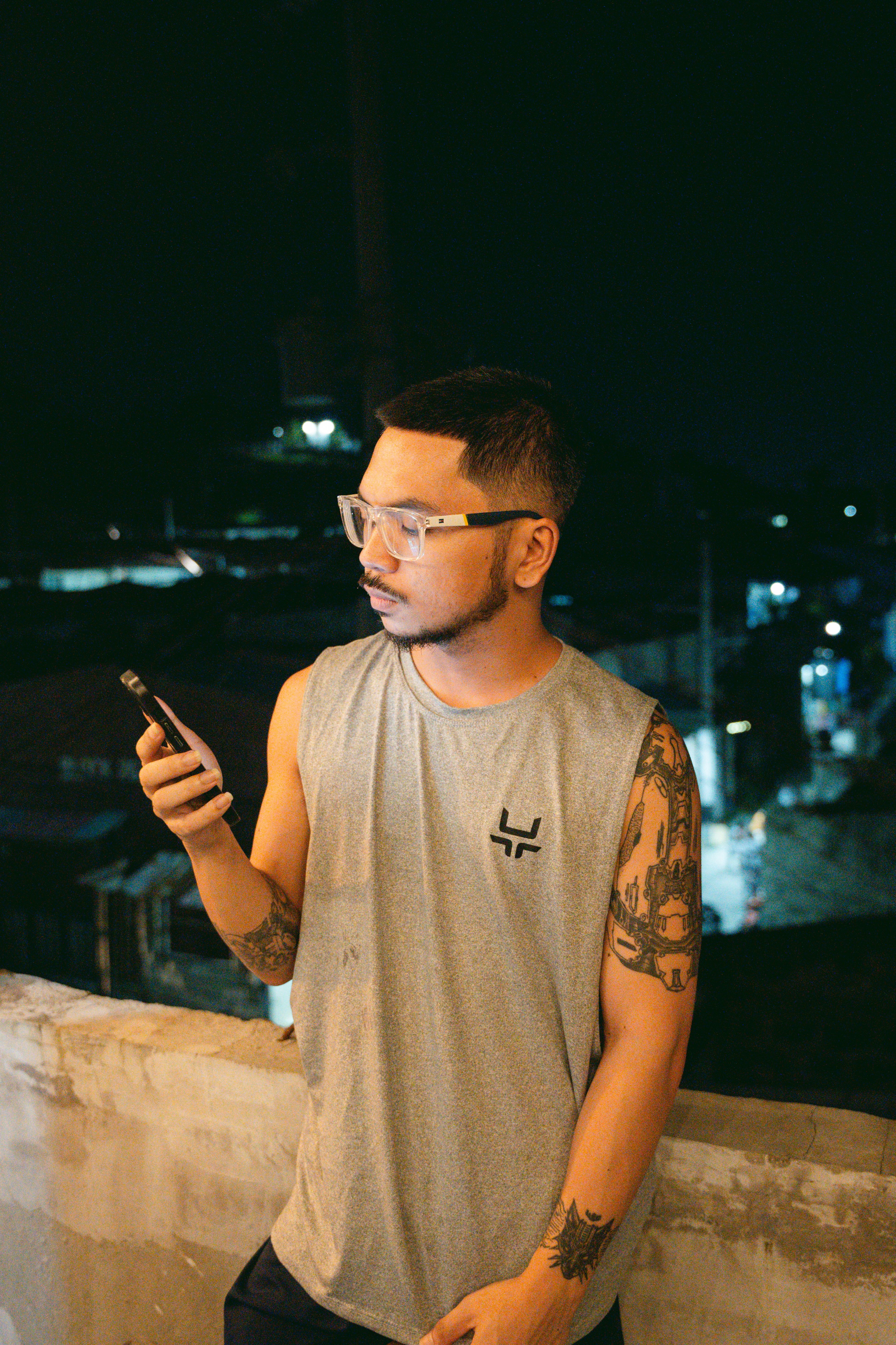 A tattooed man in casual wear using a smartphone on a balcony at night.