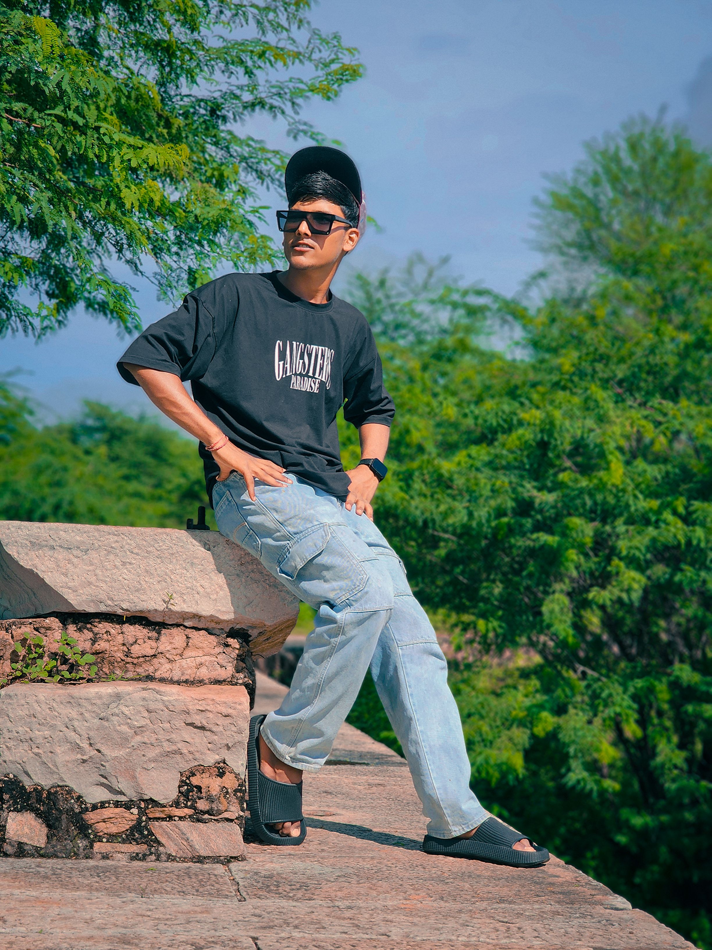 Stylish man sitting on a stone wall outdoors in casual fashion with sunglasses and cap.