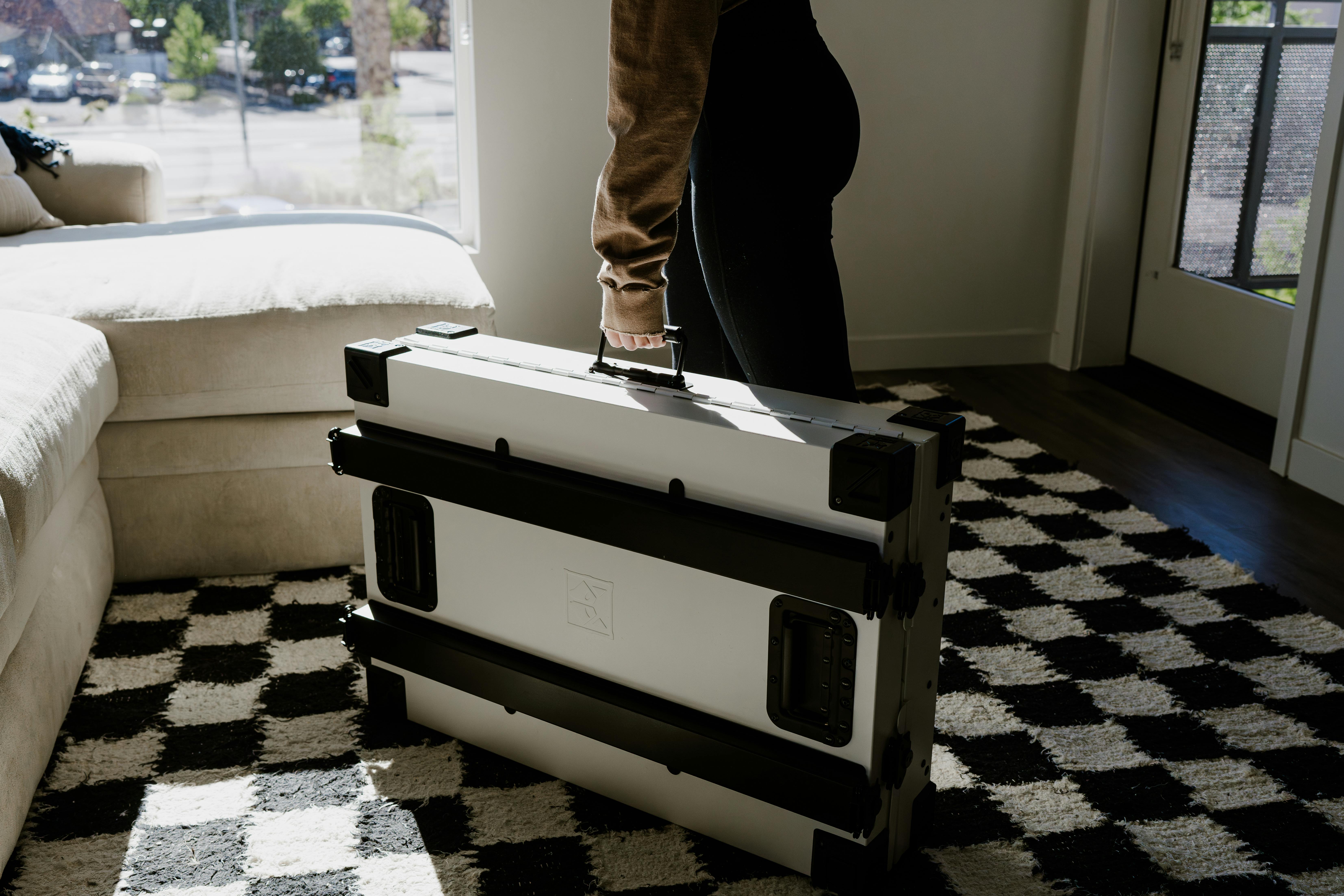 Woman holding a portable military-grade dog crate in a sunlit living room setting.