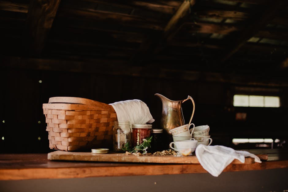 A rustic kitchen scene featuring a basket, brass jug, and herbs on a wooden table.