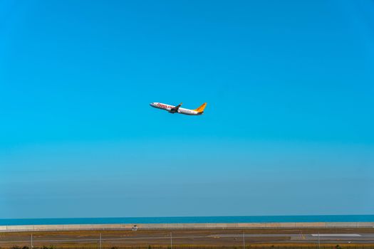 An airplane takes off from an airport runway, soaring into the clear blue sky over the ocean.