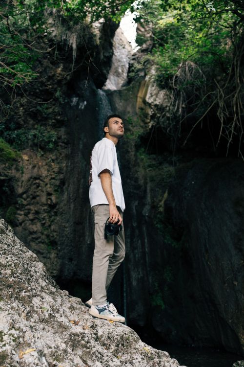 Free A man holding a camera stands by a waterfall in Merzifon, Amasya, Türkiye, surrounded by rocky terrain and lush greenery. Stock Photo