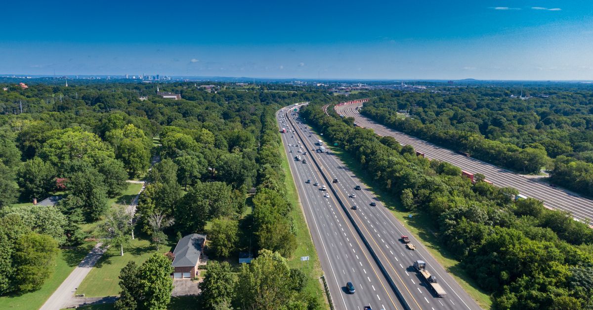 Aerial drone shot of a highway surrounded by trees, leading towards a distant city skyline under a bright blue sky.