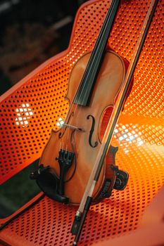 Close-up of a violin resting on an orange perforated chair, enhanced with warm lighting.